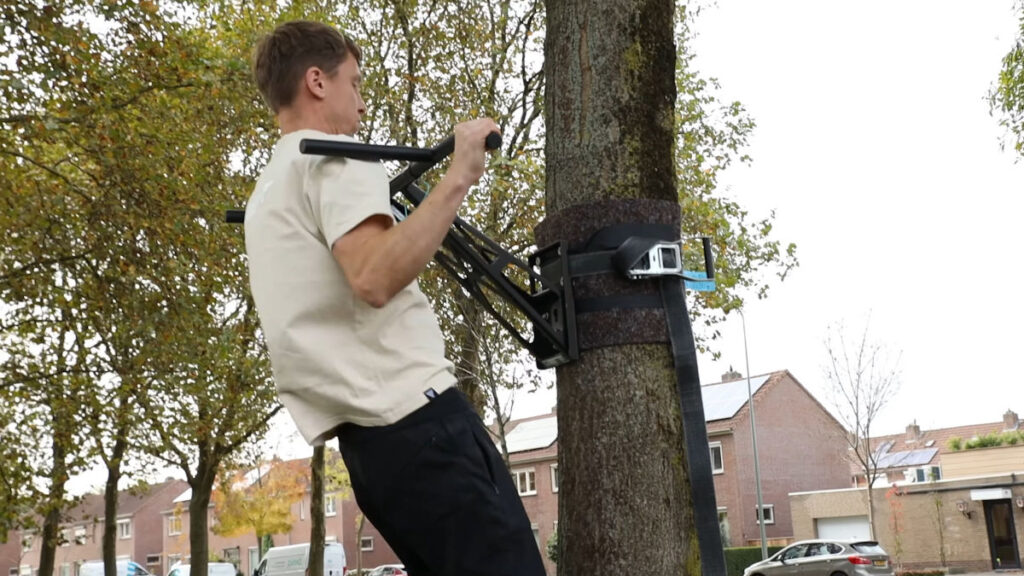 Man in white shirt performing a pull up on the Mobile Pullup and Dip Bar mounted on a tree outside with a row of houses in the background.