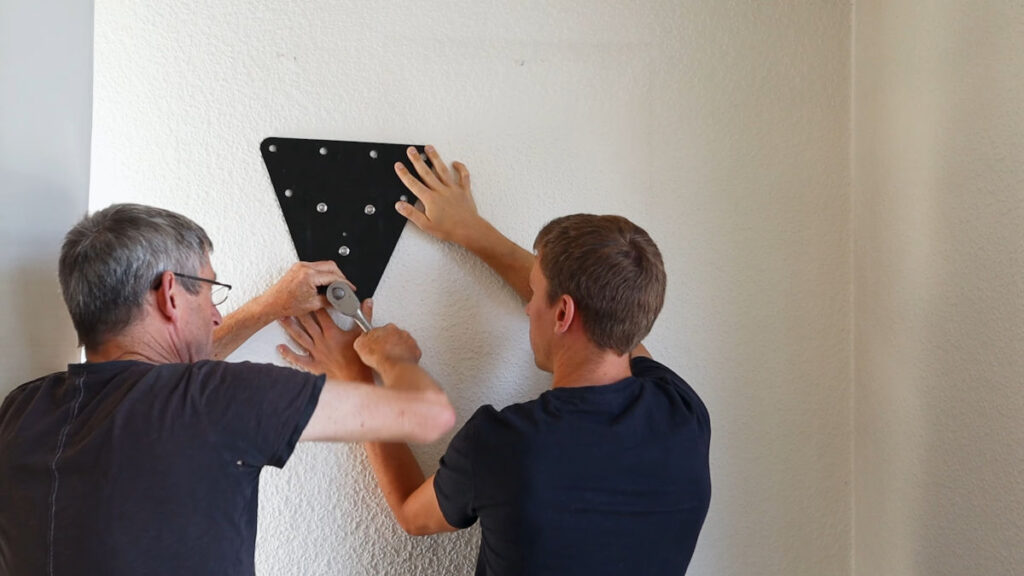 Two men holding a trapezoid base plate against the wall with the man on the left fastening a bolt that attaches the base plate, during installation of the Pullup and Dip Bar.