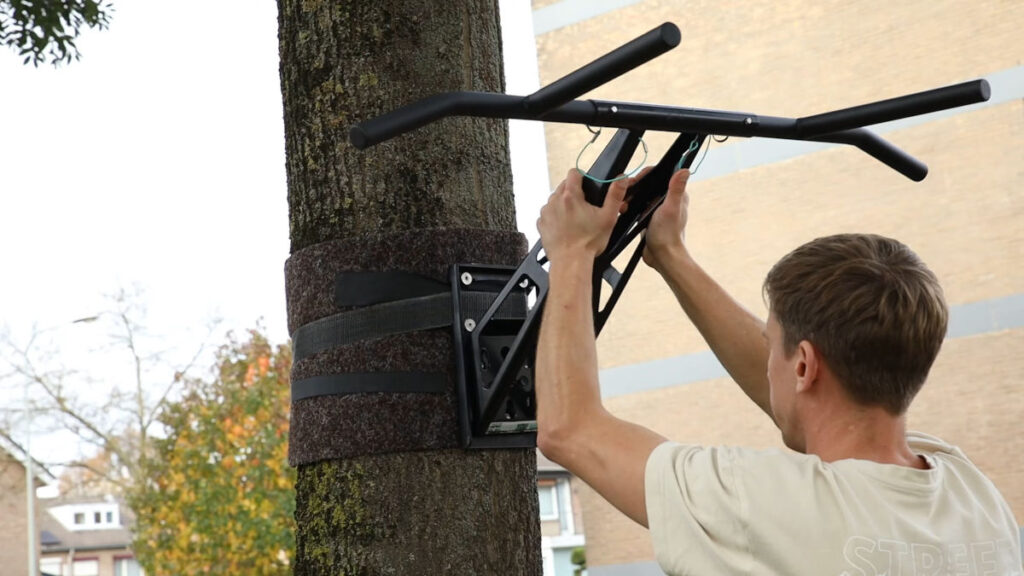 Man in white shirt applying the Mobile Pullup and Dip Bar to its tree mount outside with houses in the background.