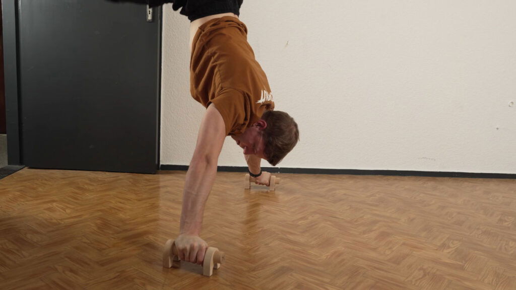 Man in brown shirt peforming a wide handstand on the Kensui Fitness YinYang Pro parallettes.