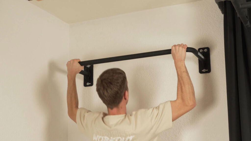 Man in white shirt perfoming a wide Pull-Up on the Gornation Pull-Up Bar "Multi" mounted on a white wall.