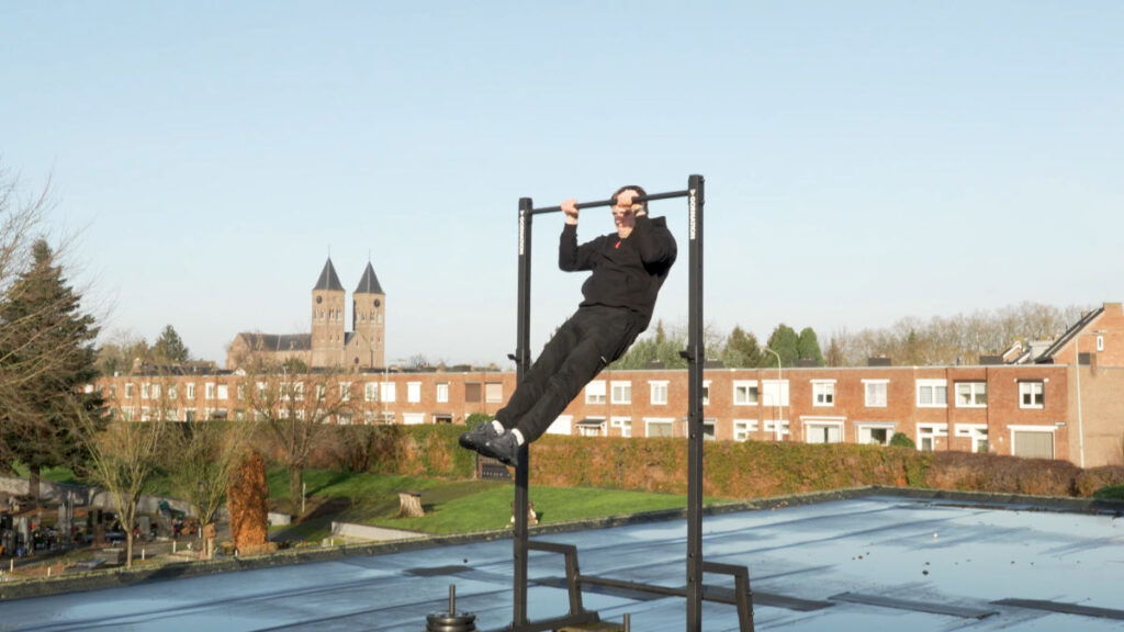 Man in black hooding performing a Pull-Up on the Gornation Pull-Up Station, the Gornation Pull-Up Bar standing outside on a roof with a neo-gothic church in the distant background.