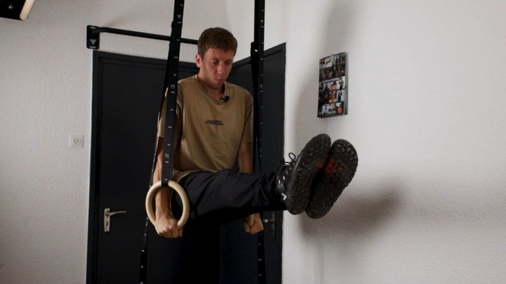 Man in beige shirt and black jogging trousers performing an L-Sit on a pair of Gymnastic Rings.