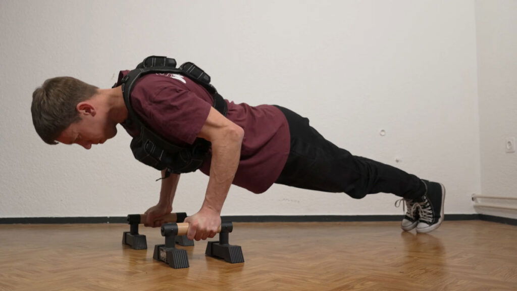 Man in red shirt doing push ups while wearing the Gravity Fitness Weighted Vest of 20kg with the assistance of a pair of Gravity Fitness parallettes.