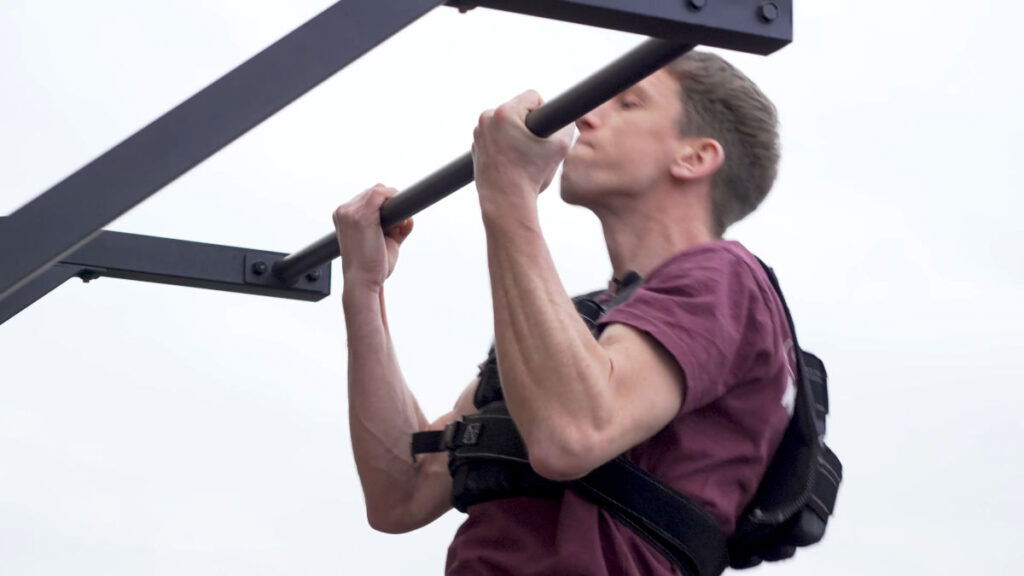 Man in red shirt doing chin ups outside while wearing the Gravity Fitness Weighted Vest of 20kg.