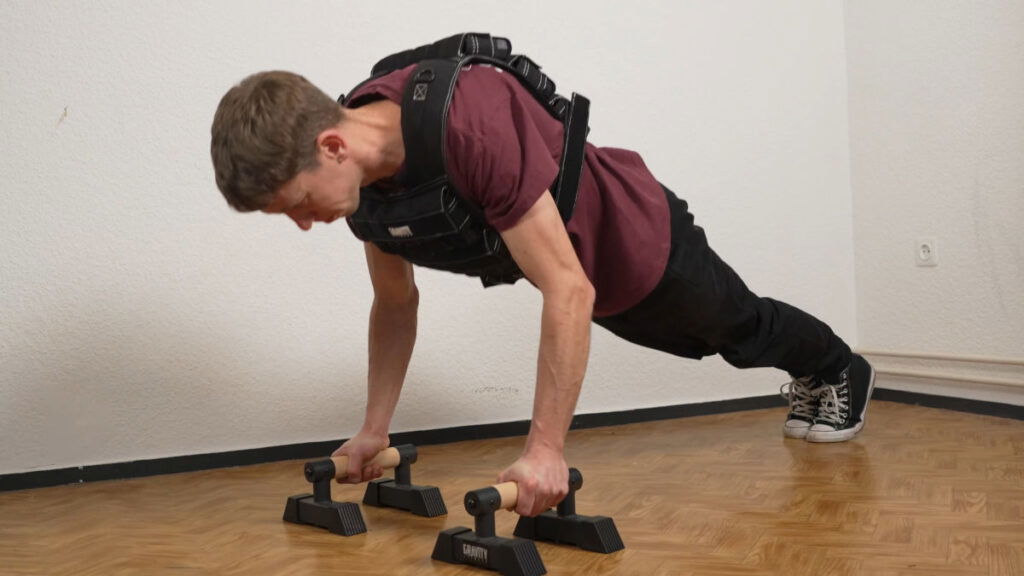 Man in red shirt doing push ups while wearing the Gravity Fitness Weighted Vest of 30kg with the assistance of a pair of Gravity Fitness parallettes.