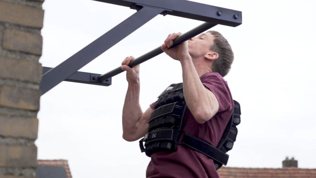 Man in red shirt doing pull ups outside while wearing the Gravity Fitness Weighted Vest of 30kg.