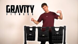 Man in wine red shirt standing behind and pointing at the three Gravity Fitness Weighted Vests on a dip bar. The Gravity Fitness logo is prominently displayed beside him.