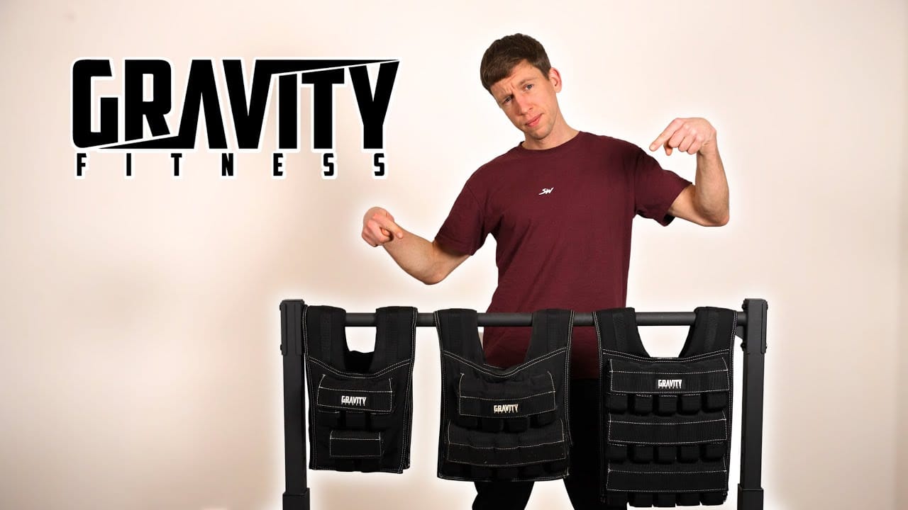 Man in wine red shirt standing behind and pointing at the three Gravity Fitness Weighted Vests on a dip bar. The Gravity Fitness logo is prominently displayed beside him.