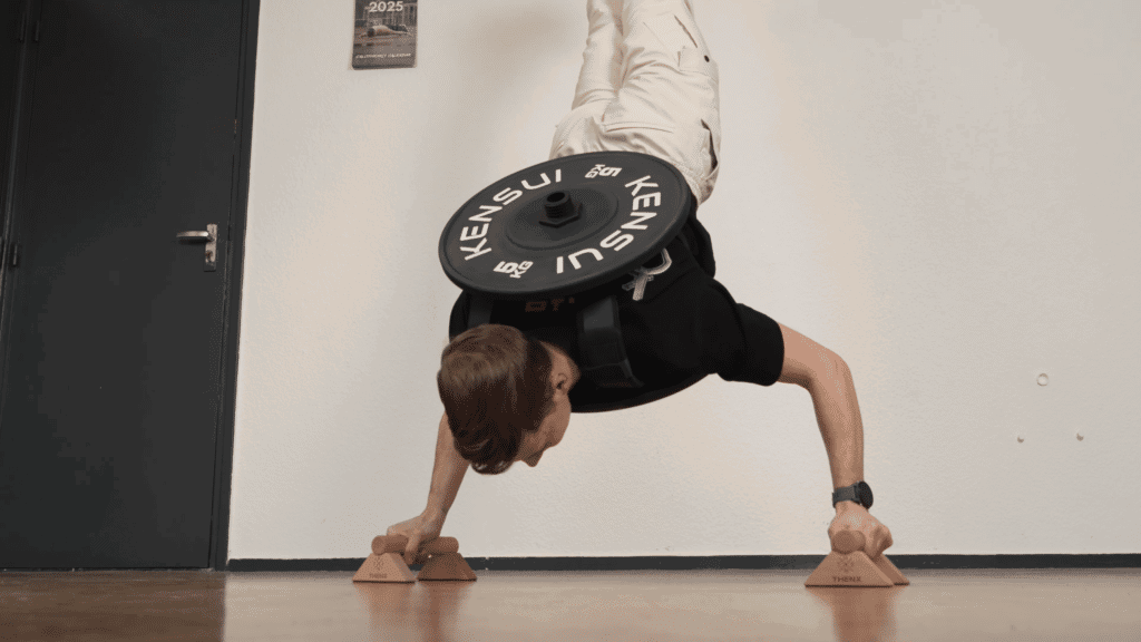 Man doing a handstand push up wearing a Kensui EZ vest plate loaded weight vest with the assistance of a pair of ThenX push up bars.