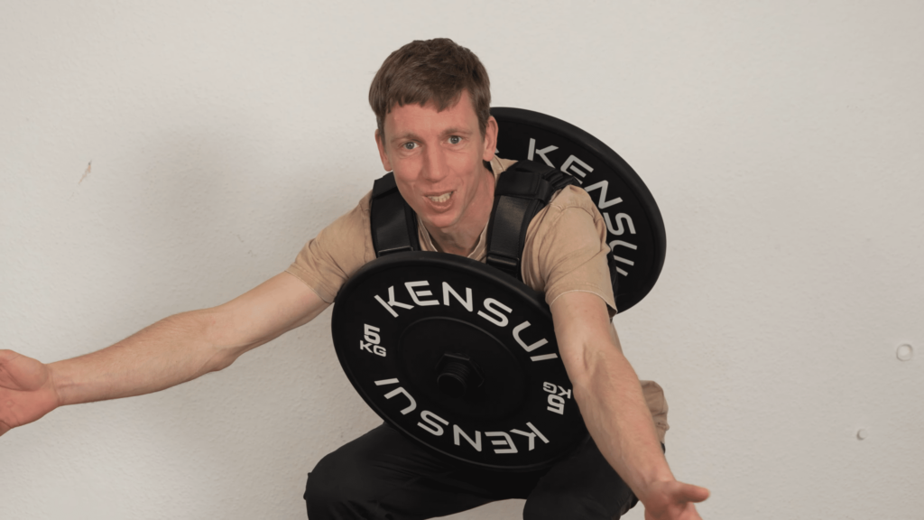 Man smirking in contempt as he invites the viewer to see how the plate of his plate loaded weight vest obstructs his movement while doing squats.