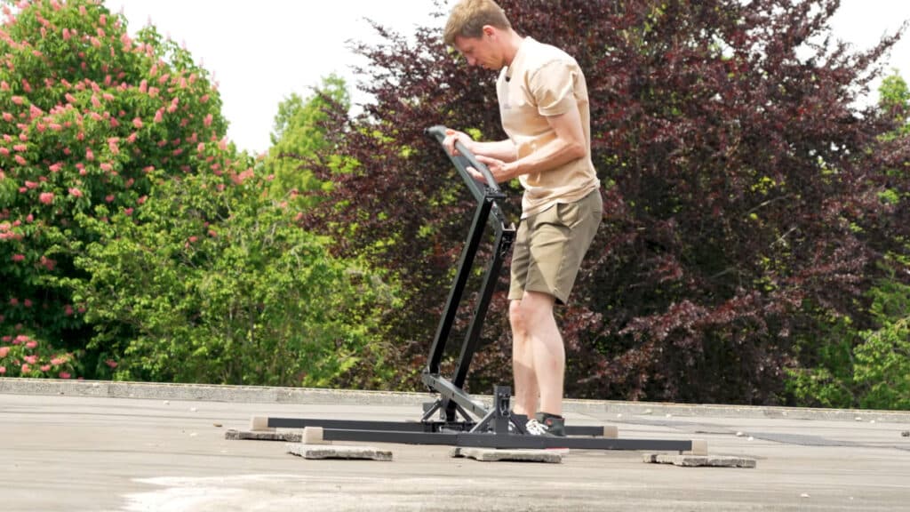 Man in beige shirt unfolding the foldable BullBar Free-Standing Pull-Up Bar on a roof with trees in the background.