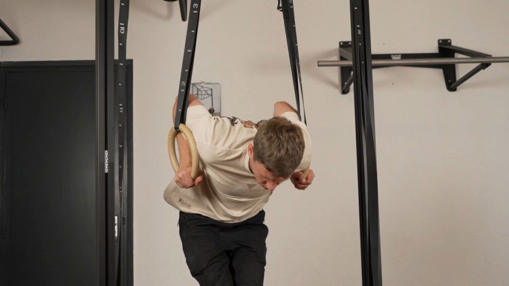 Man in beige shirt performing a Ring Muscle Up on a set of gymnastic rings suspended from a FitnessKit Basic Strength Bar.