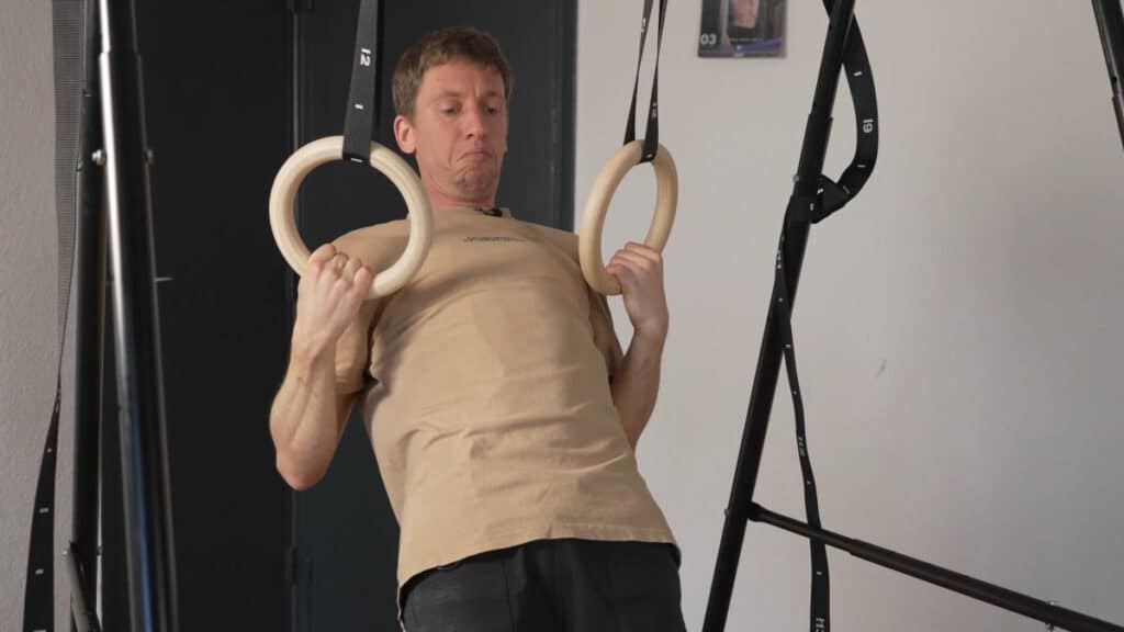 Man in beige shirt completing a body row using Gymnastic Rings suspended from a Khanh Trinh Pull-Up Bar.