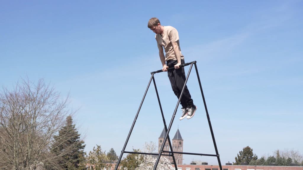 Man in beige shirt completing a Muscle-Up on the Khanh Trinh Pull-Up Bar standing outside on a roof with a large church in the background.