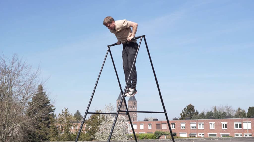 Man in beige shirt performing a Muscle-Up on the Khanh Trinh Pull-Up Bar outdoors on a roof with a large church in the background.