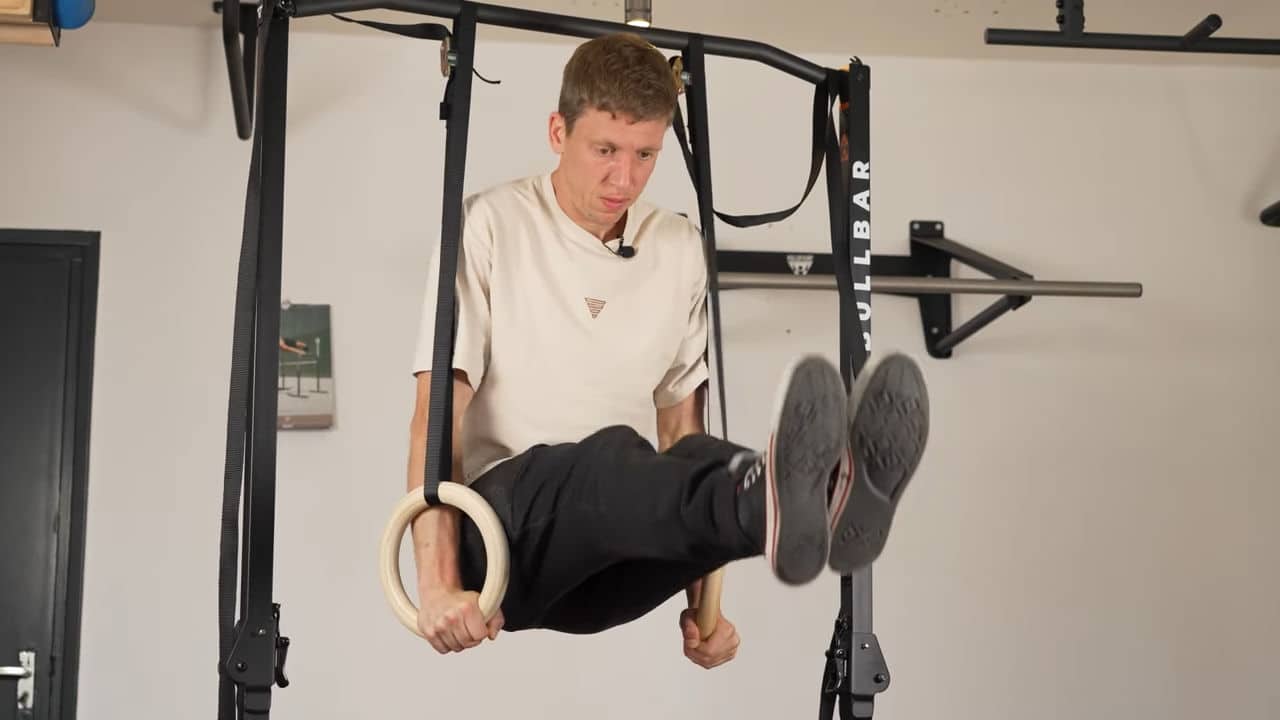 Man in beige shirt doing an L-sit on a pair of gymnastic rings suspended from a bullbar 2.0 foldable pull-up bar.