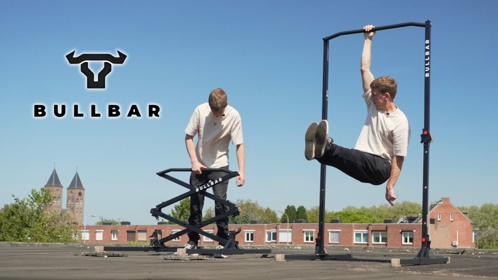 Man in beige shirt setting up and hanging with one arm from the Bullbar 2.0 foldable pull-up bar on a roof with a church in the background.