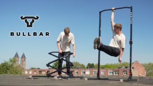 Man in beige shirt setting up and hanging with one arm from the Bullbar 2.0 foldable pull-up bar on a roof with a church in the background.