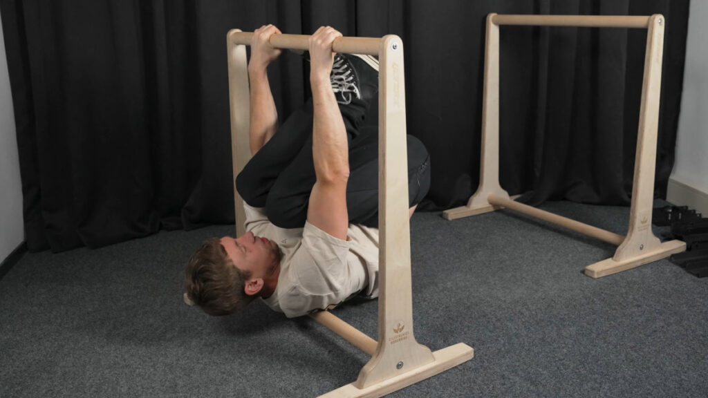 Man in white shirt attempting a skin the can on the gorletic wooden dip bars but failing.