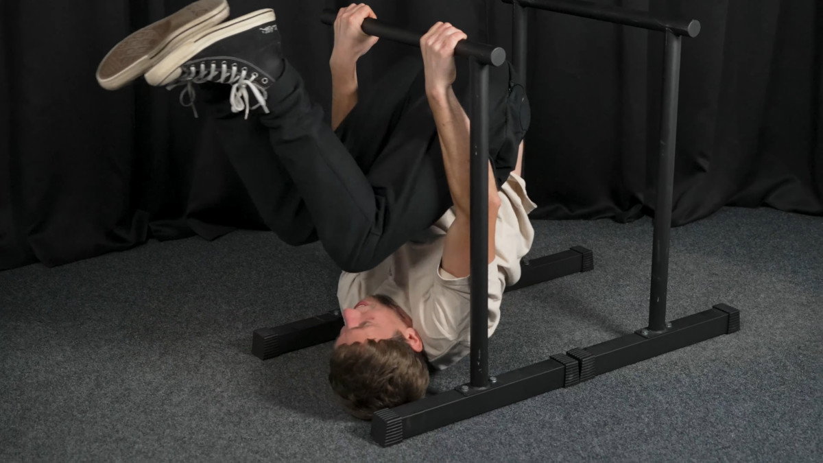 Man in white shirt attempting a skin the cat on the ramass dip bars but failing because it is too low.