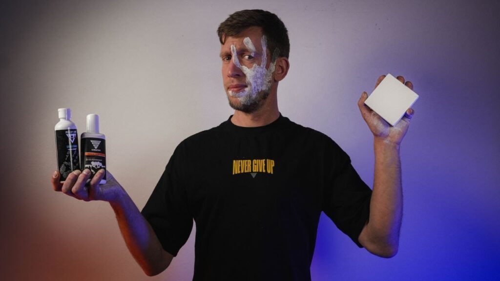 Man with a white handprint on his face holding two bottles of liquid chalk and a block of dry chalk in his hands, looking doubtfully at the camera.