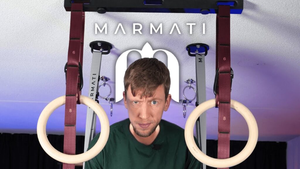 Man in green shirt standing between two marmati gymnastic rings suspended from red straps and looking doubtfully at the camera.