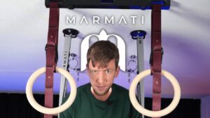 Man in green shirt standing between two marmati gymnastic rings suspended from red straps and looking doubtfully at the camera.