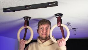 Man hanging on to a pair of gymnastic rings that are fastened to the ceiling by ceiling hooks, and looking doubtfully at the camera.