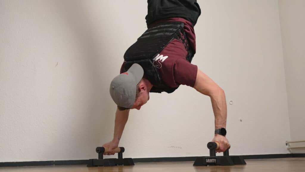 Man in red shirt and wearing a baseball cap doing handstand push-ups on the gravity fitness parallettes and wearing the force fitness weighted vest.