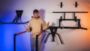 Man in beige hoodie standing between multiple wall-mounted dip bars and pointing his thumbs at them, looking questioningly at the camera.