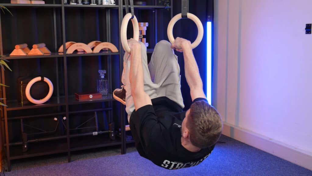 Man doing a front lever on a pair of gravity fitness gymnastic rings with multiple other wooden calisthenics equipment tools in the background.
