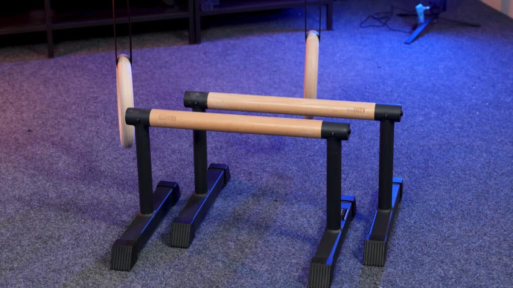 A pair of gravity fitness medium parallettes standing in front of a pair of gravity fitness gymnastic rings as part of a complete, minimalist home gym.