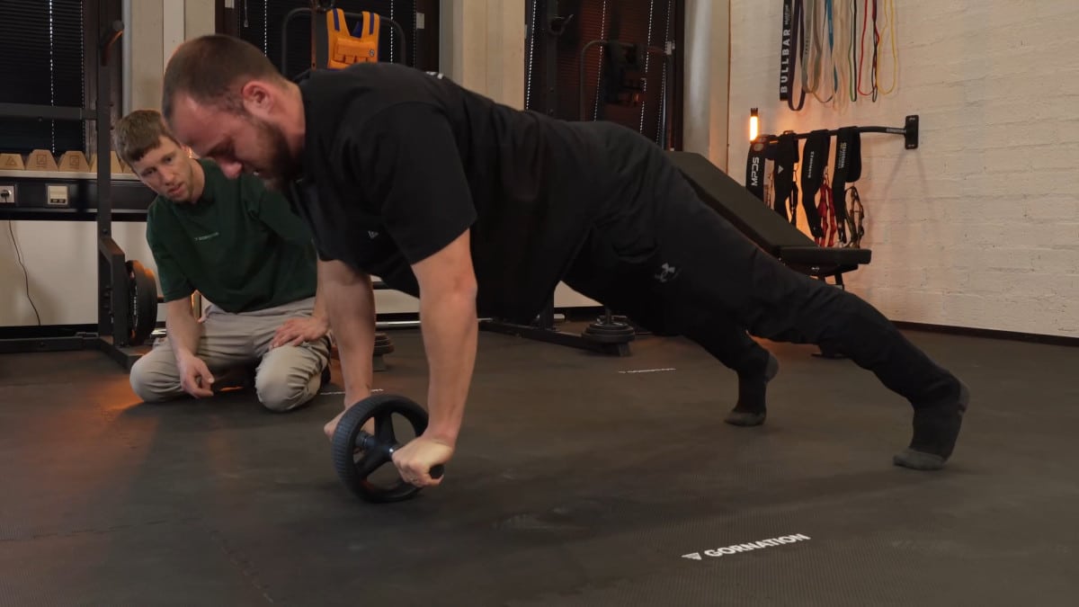 Man in black sports attire doing push ups on the rogue ab wheel with another man monitoring his movements in the background.