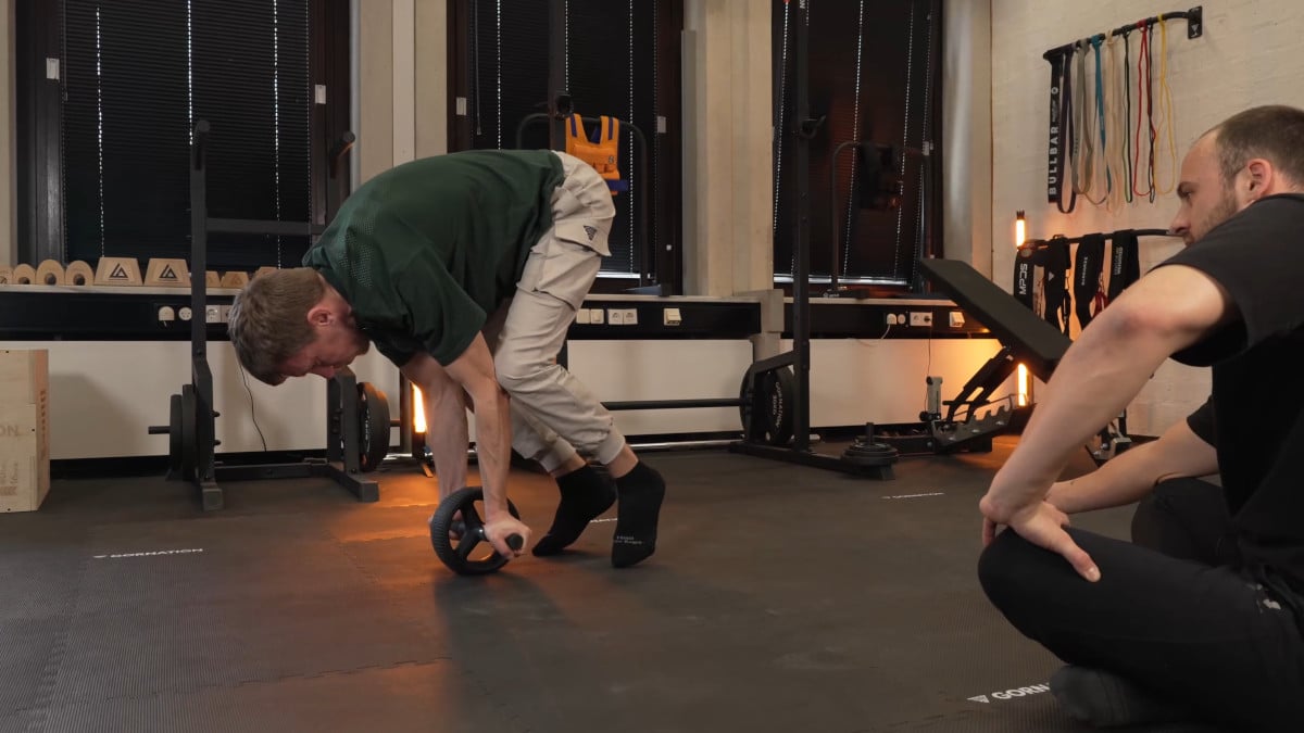 Man in green shirt attemping to balance on the rogue ab wheel by lifting his feet off the floor with another man observing him from the side.