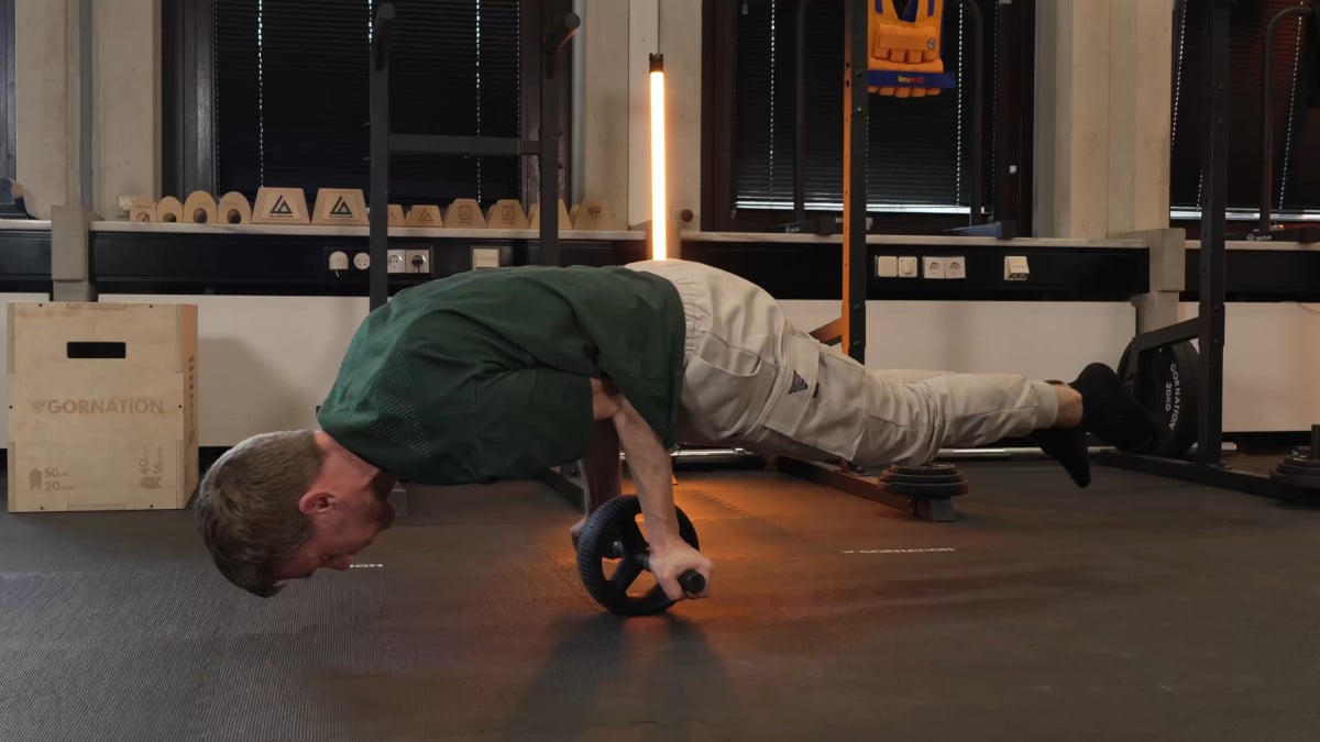Man in green shirt doing a planche balancing on the rogue ab wheel in a gym setting.