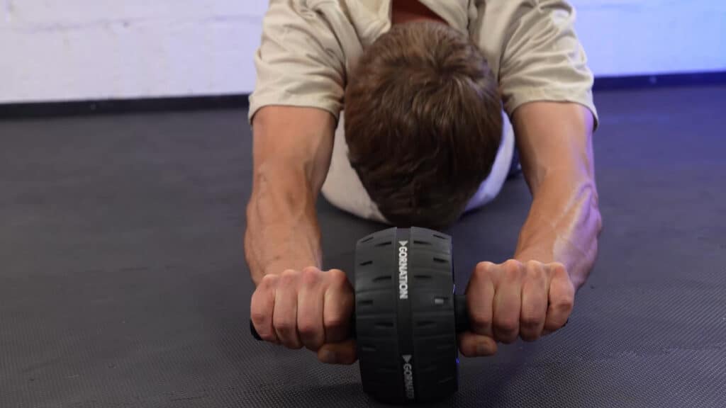 Front image of a man doing ab rollouts, pushing a gornation ab roller towards the viewer over a gym floor.