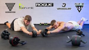 Two athletic men doing ab rollouts on an assortment of ab rollers from various brands while looking questioningly at the viewer.