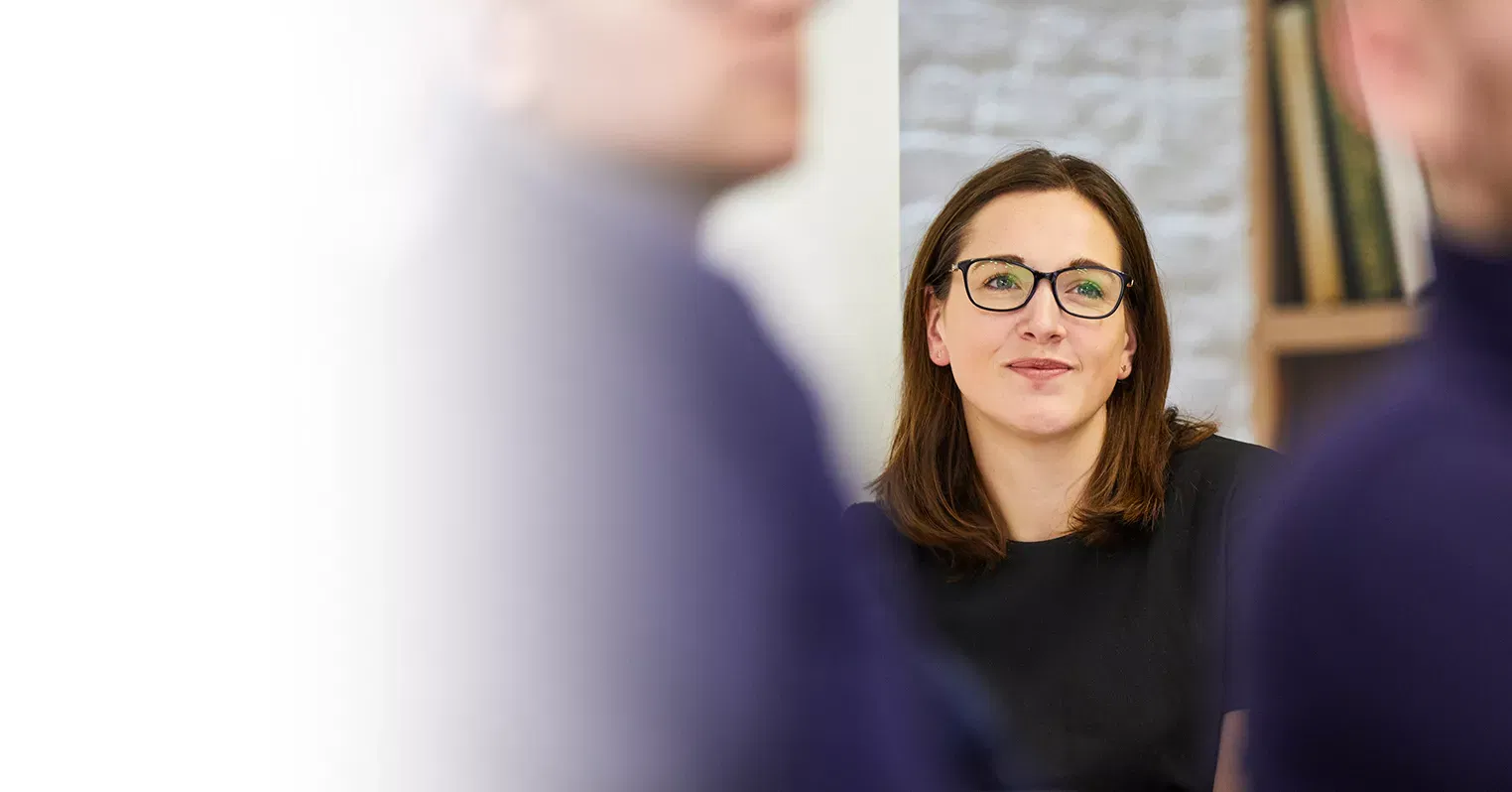 A woman wearing glasses, among a couple of out-of-focus people