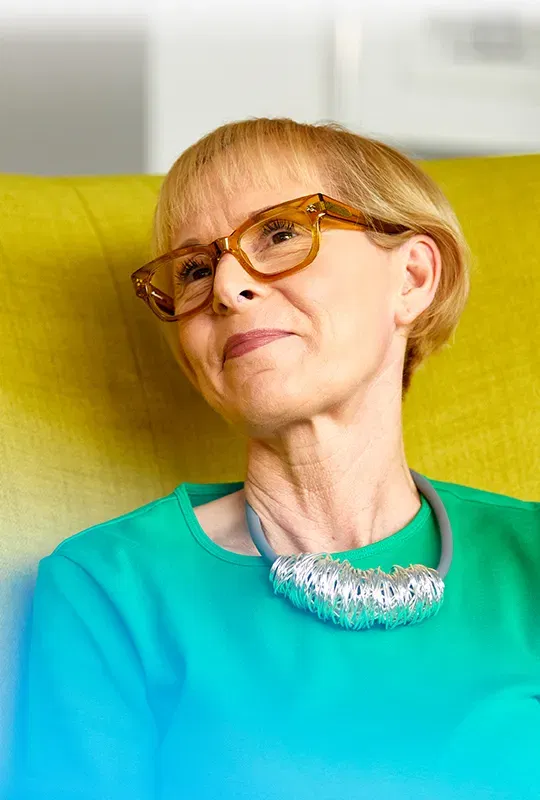 A woman in glasses and a necklace sits in a green wing-backed chair