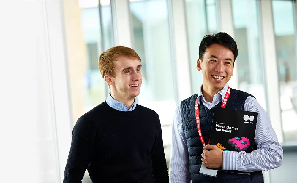 Photo of two smiling men, the one on the right is weating a lanyard and holding a copy of an Oury Clark guide to video games tax relief