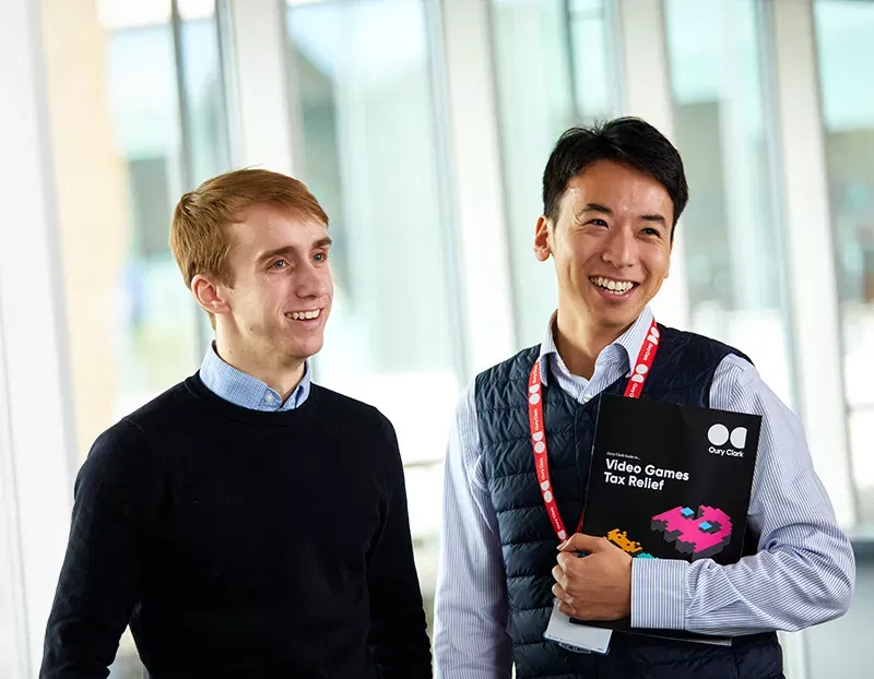 Photo of two smiling men, the one on the right is weating a lanyard and holding a copy of an Oury Clark guide to video games tax relief