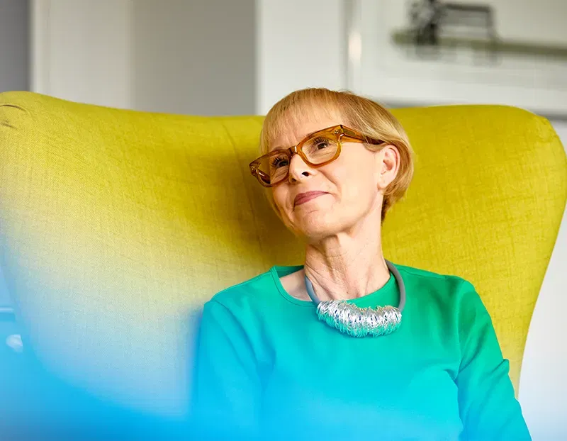 A woman in glasses and a necklace sits in a green wing-backed chair