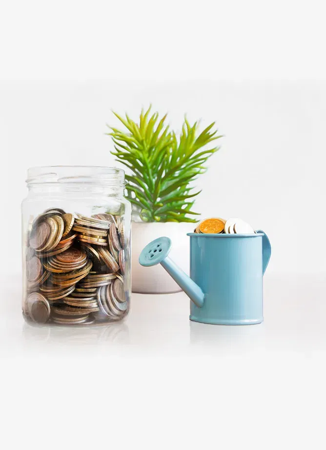 A glass jar full of coins stands next to a miniature watering can full of coins. A plant is in the background