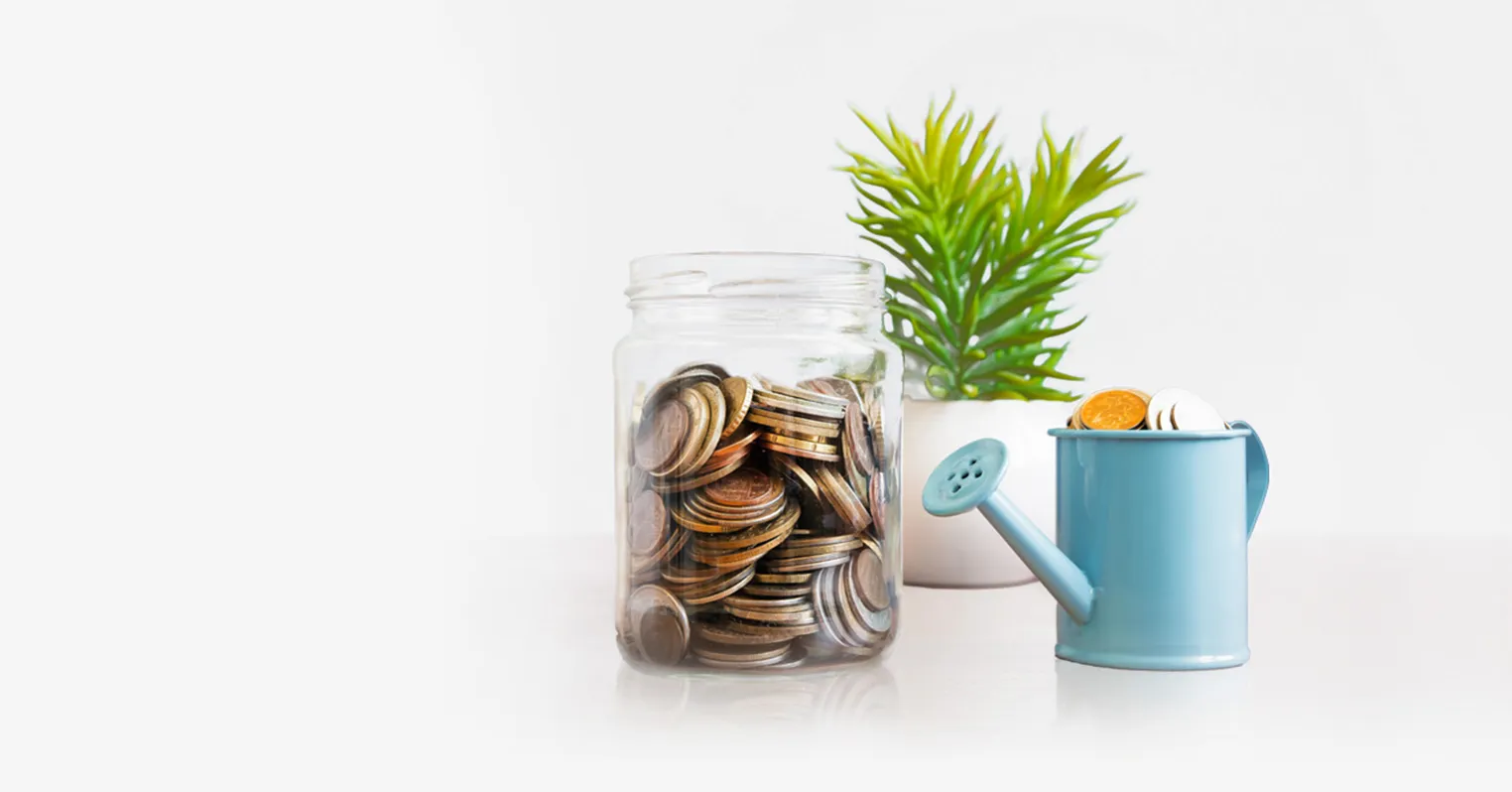 A glass jar full of coins stands next to a miniature watering can full of coins. A plant is in the background