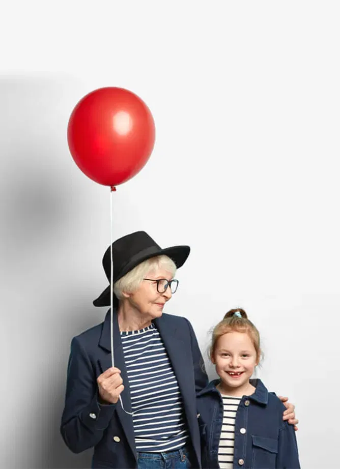 Photo of an older woman wearing a hat and holding a red balloon, with her arm around a little girl who smiles at the camera