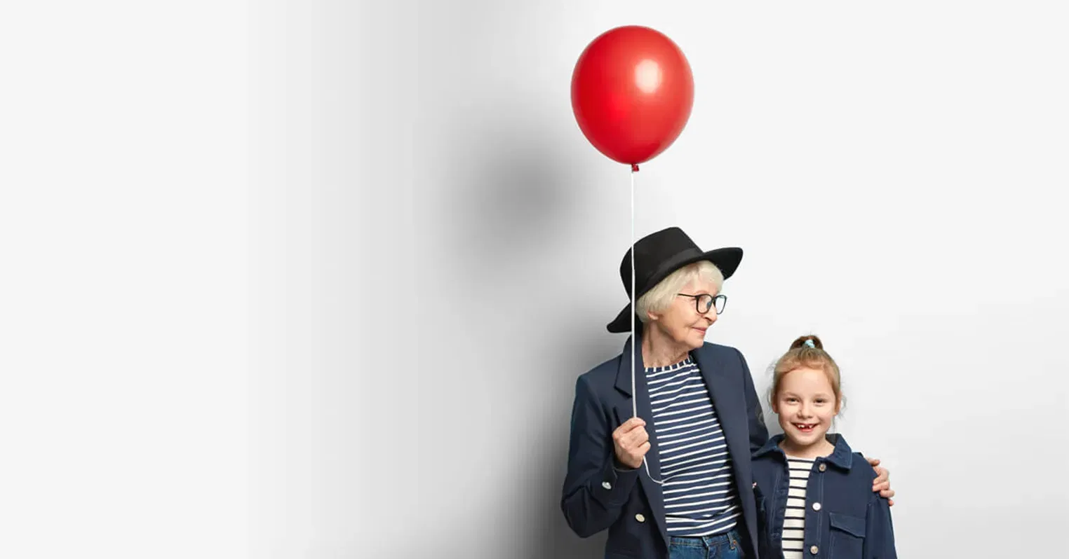 Photo of an older woman wearing a hat and holding a red balloon, with her arm around a little girl who smiles at the camera