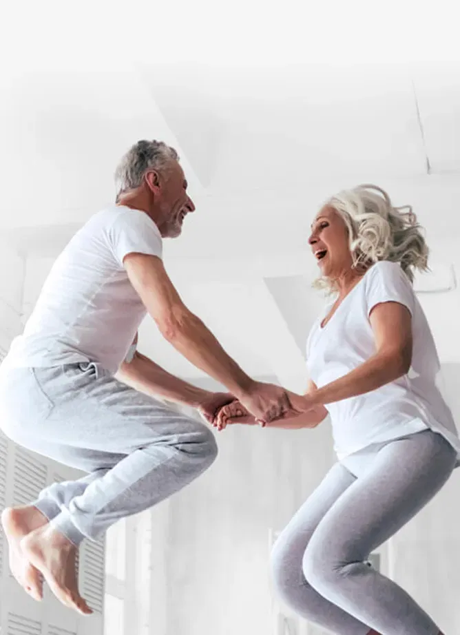 Photo of a man and woman wearing grey jogging pants and white t-shirts jumping up into the air and laughing