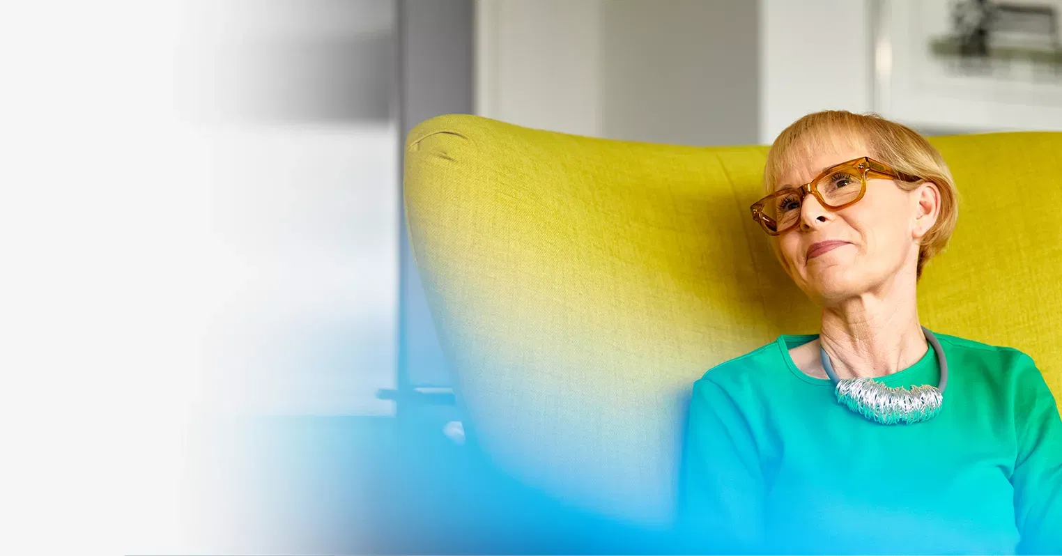 A woman in glasses and a necklace sits in a green wing-backed chair