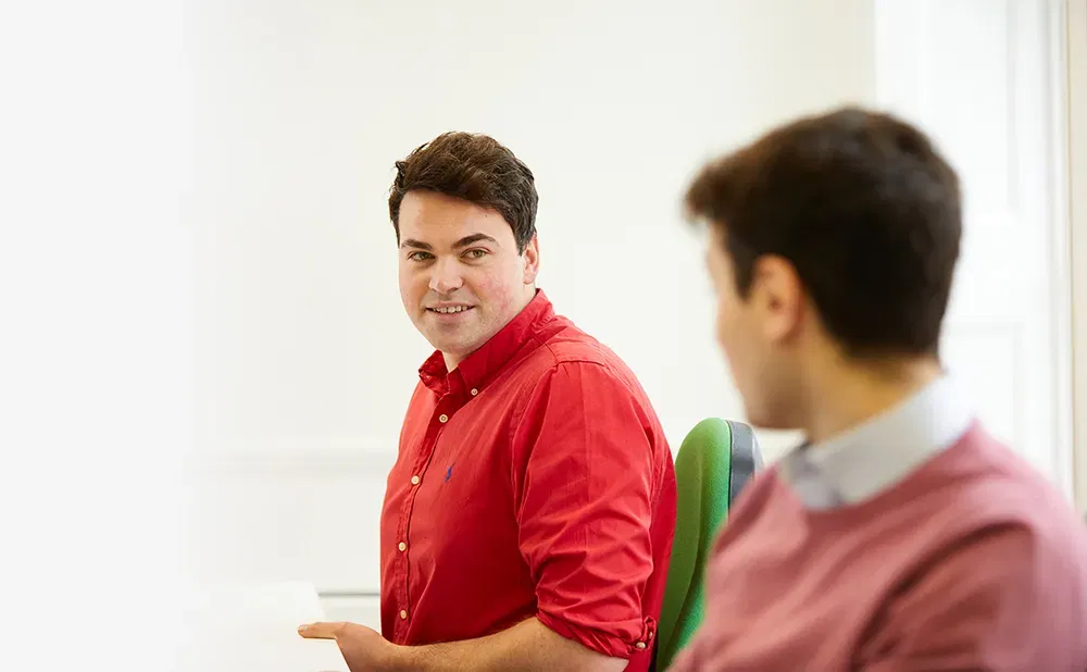 A man in a red shirt looks at an out of focus man in a pink jumper